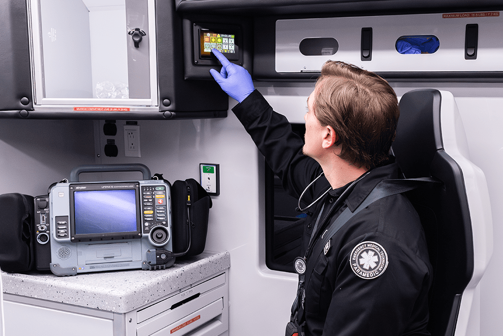 Paramedic seated in the patient module adjusting the Demers ambulance multiplex control touchscreen, with a cardiac monitor and organized cabinetry visible in the background