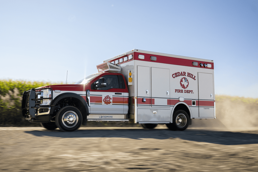 Side view of a Demers Type I ambulance driving on a rural road, with dust rising behind it.