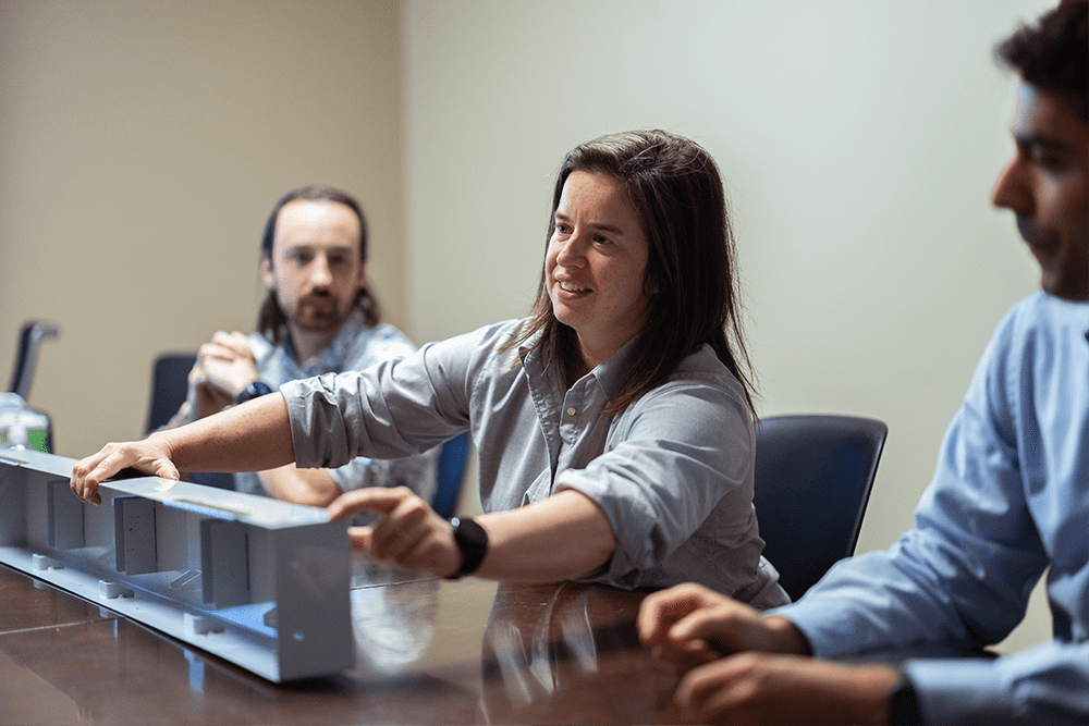 Group of Demers employees seated at a conference table, with one person presenting and pointing to a scale model during a meeting